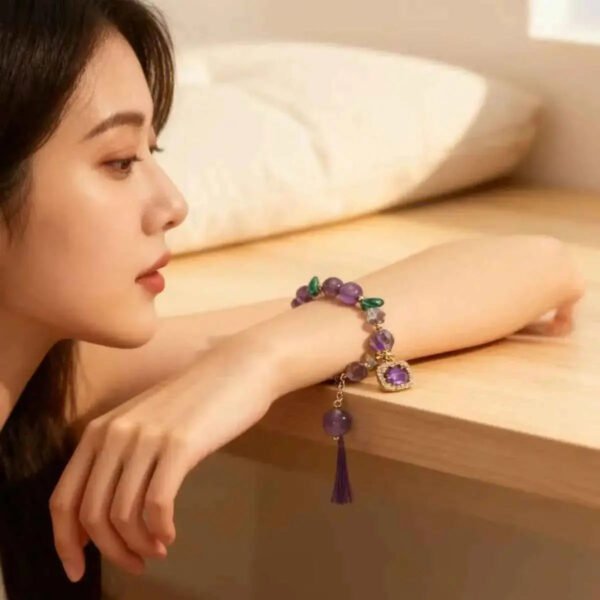 Amethyst gemstone bracelet on wrist with purple beads, gold chain, green accents, and dangling charm.